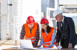 OSP 1 A group of business people are having a meeting on an oil industry site. The two engineers (a man and a woman) wearing reflective clothing and red working helmets are having a discussion and planning something with a senior investor who is wearing an elegant black suit and white helmet. They are watching something on a white laptop placed on a brown table in front of them together with some plans. In the background we can see the rafinery.
