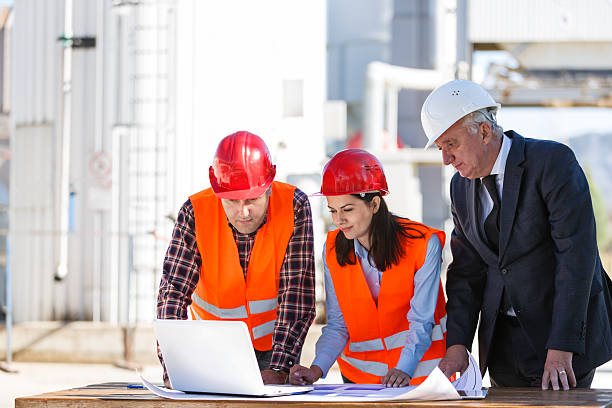 A group of business people are having a meeting on an oil industry site. The two engineers (a man and a woman) wearing reflective clothing and red working helmets are having a discussion and planning something with a senior investor who is wearing an elegant black suit and white helmet. They are watching something on a white laptop placed on a brown table in front of them together with some plans. In the background we can see the rafinery.